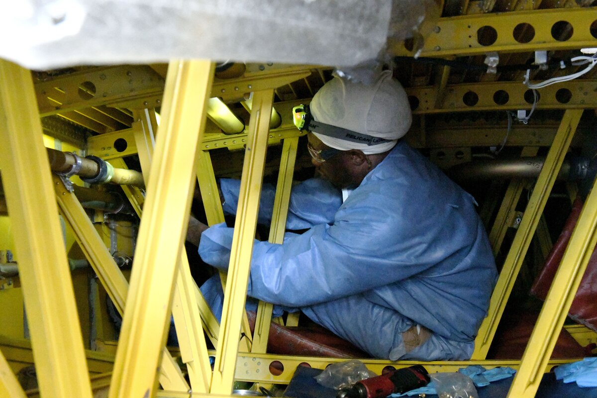 A man in blue protective gear is crouching in a tight area surrounded by yellow metal struts.