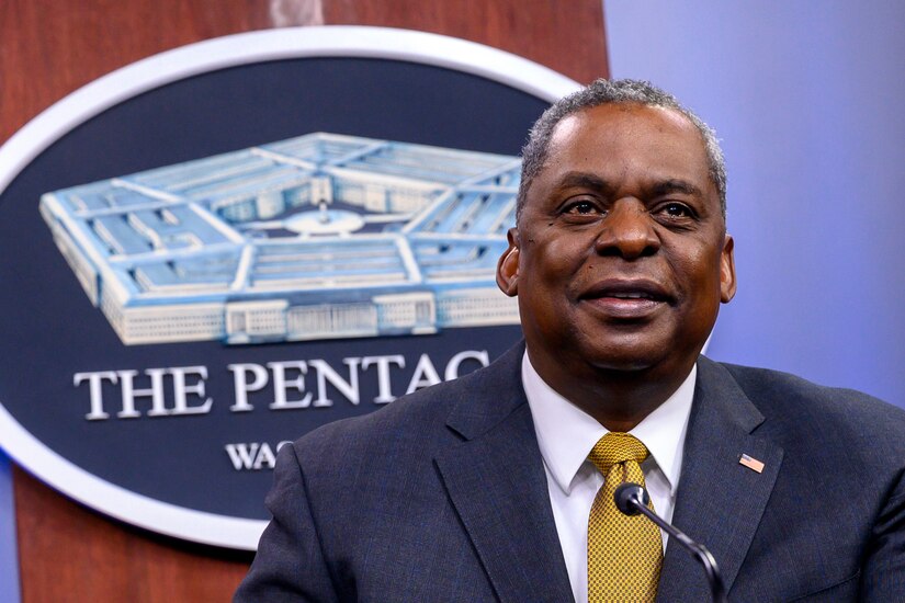 A man wearing a business suit sits at a table and speaks into a microphone.