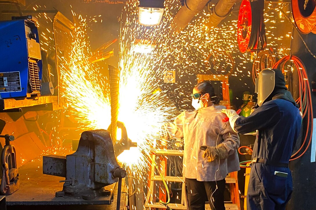 Sparks fly as two sailors operate a cutting tool in an industrial-type work area.