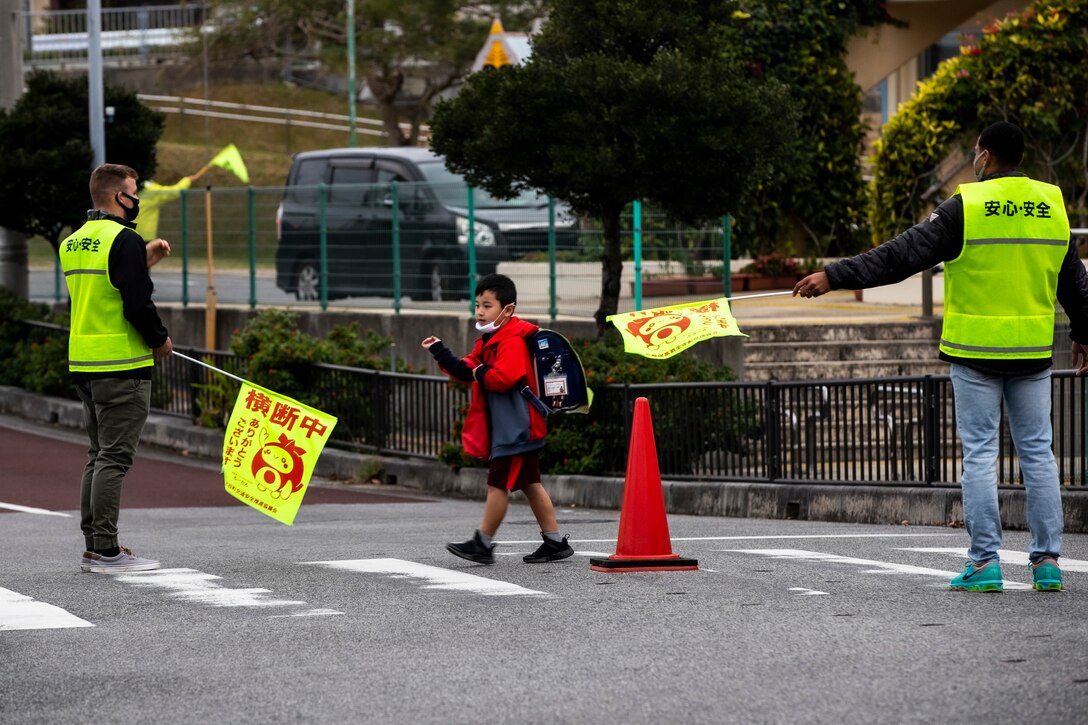 U.S. Marine Corps Lance Cpl. Kyle Knepper, left, and Lance Cpl. James Cruz, right, heavy equipment operators with 9th Engineer Support Battalion, 3rd Marine Logistics Group, act as crossing guards while a student crosses a crosswalk at Kitatama Elementary School, Chatan, Okinawa, Japan Jan. 7, 2021. Every Tuesday and Thursday, military community volunteers serve as crossing guards to assist elementary students and staff crossing a busy neighborhood intersection. The volunteers received letters of appreciation for their work from the Kitatama Community Center. (U.S. Marine Corps photo by Cpl. Ryan H. Pulliam)