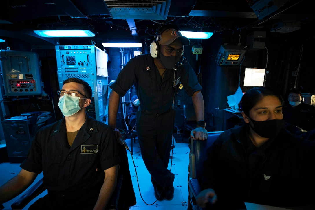 A sailor stands behind two sailors seated at a control board.