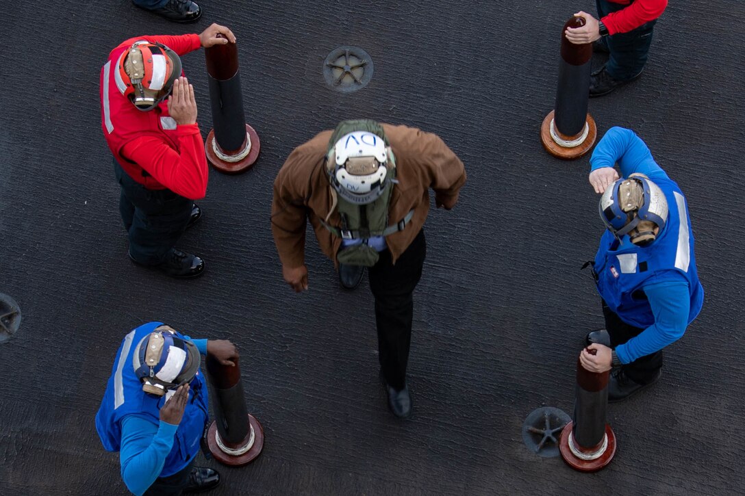 Secretary of Defense Lloyd J. Austin III, shown from overhead, walks onto a ship as sailors in red shirts and blue shirts on either side of him salute.