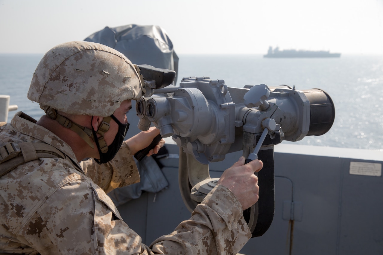 A male service member looks through a ship's bridge binoculars to scan the sea as a ship sails in the distance.