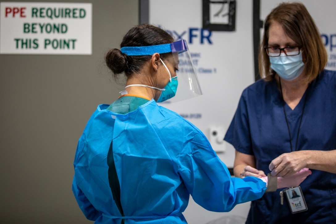 A woman aids another woman in donning personal protective equipment in a hallway while both wear face masks and medical scrubs.