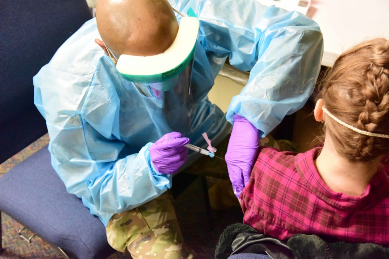 A bald man in military camouflaged pants and a medical apron with gloves and a face shield is seated to the left of a person in a red plaid shirt as he holds a needle to inoculate her against the COVID-19 virus.