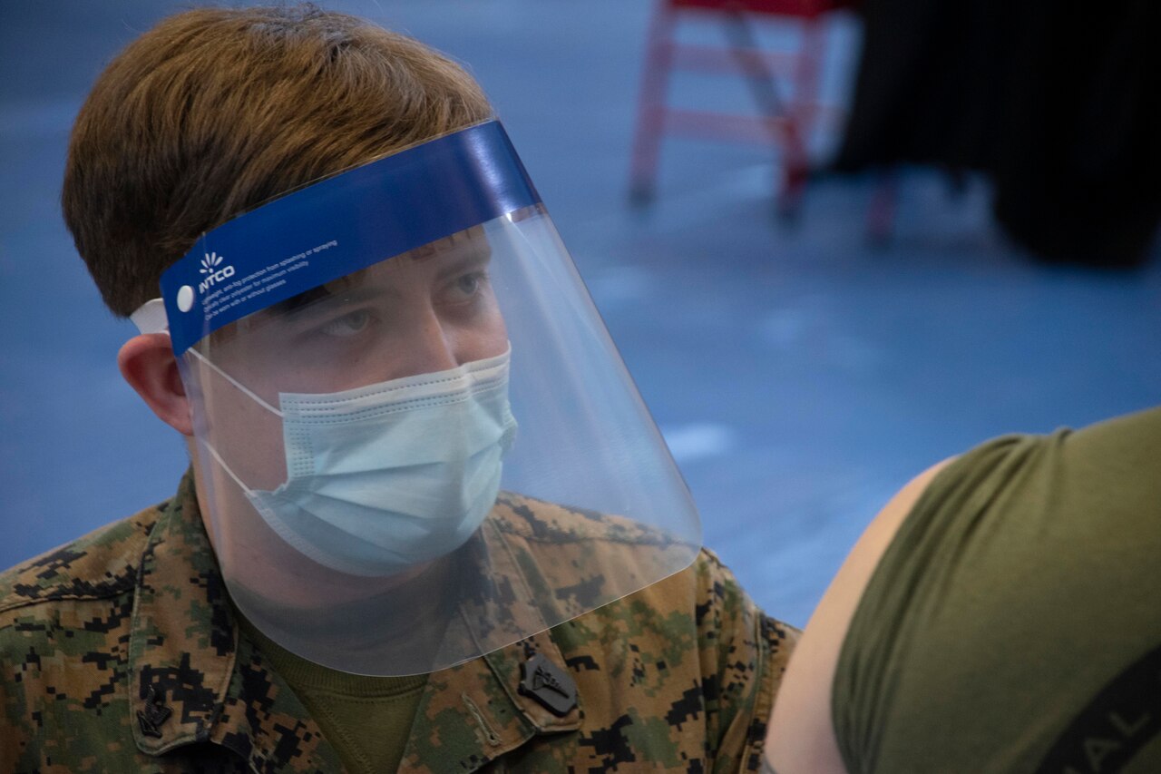 A sailor wearing a face mask and shield prepares to vaccinate a Marine.