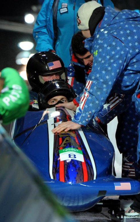 Two men wearing helmets are seated in a bobsled as others crowd around them.