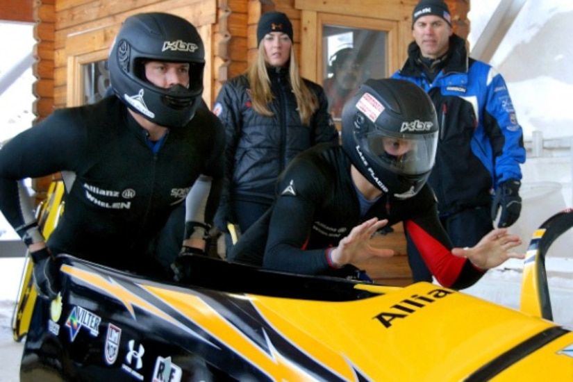 Two people wearing helmets push a bobsled as a man and woman watch.