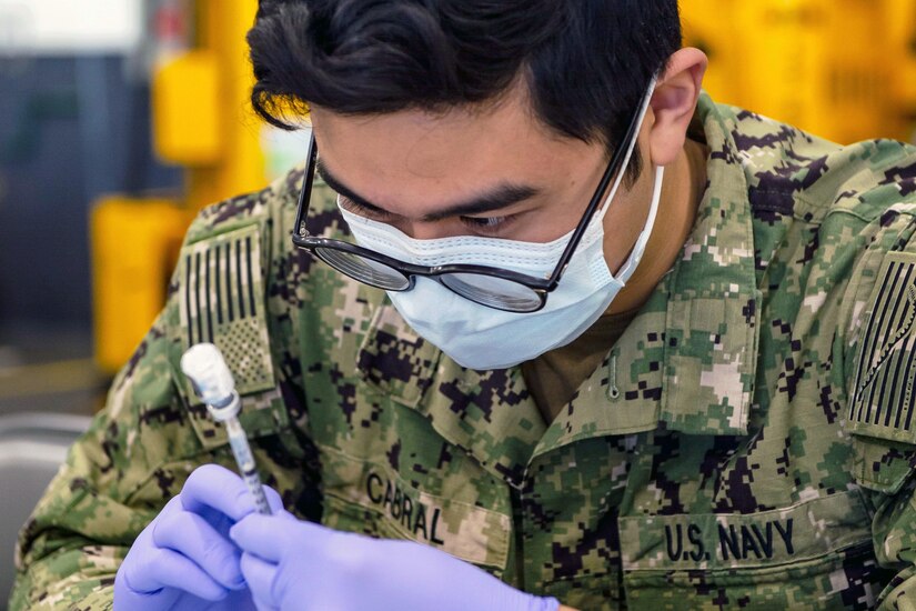 A service member wearing a face mask and gloves holds a syringe into a small bottle.