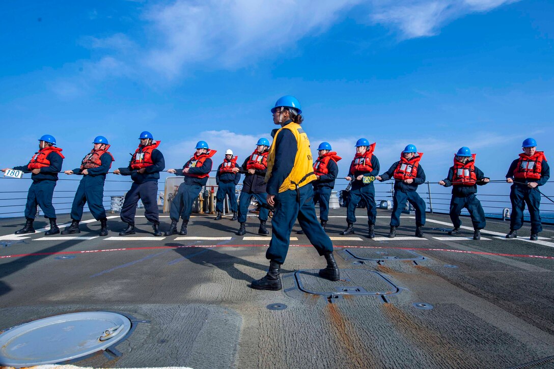 A sailor watches as a line of other sailors pull a rope.