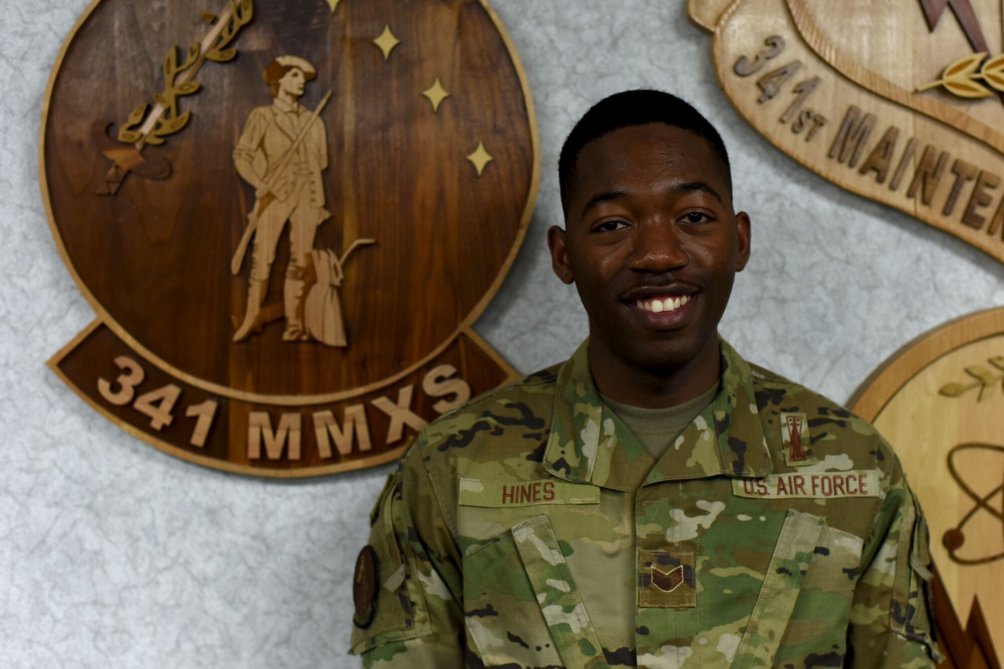 Staff Sgt. Khalif Hines stands in front of a maintenance squadron emblem and looks directly at the camera as he smiles for the photo.