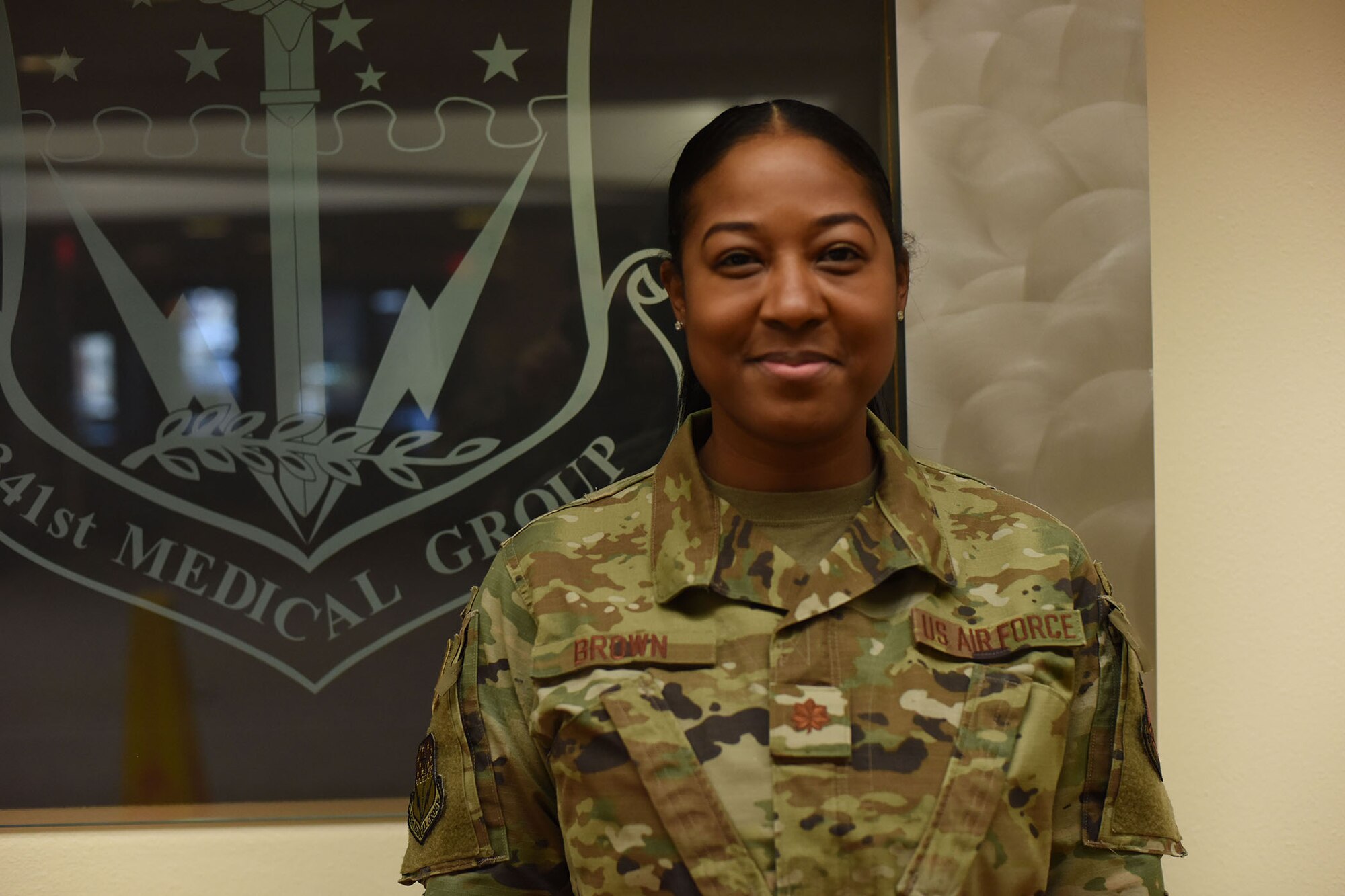 Maj. Rashida Brown stands in front of a glass wall with the 341st Medical Group emblem on it as she smiles for the photo.