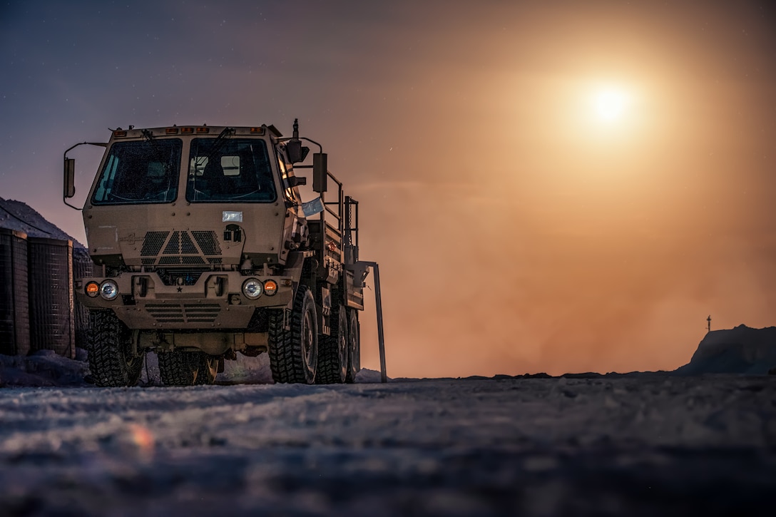 A truck sits on a dusty landscape. To the left of the truck are gravel-filled barriers.