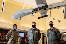 Three Airmen stand together under a hanging MQ-1 Predator aircraft and look at the camera.