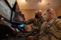 One male Airman sits in a simulator, while another male Airman points to the screens in front of them.