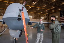 One male Airman points to the camera on an MQ-9 Reaper aircraft, while the other male Airman looks at the camera with their arms crossed.
