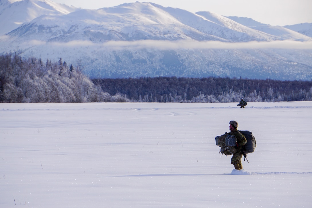 Two paratroopers walk through snow with mountains in the background.