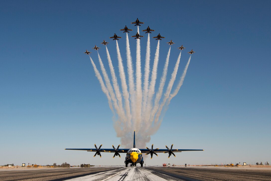 Aircraft fly in formation over an aircraft on a landing strip.