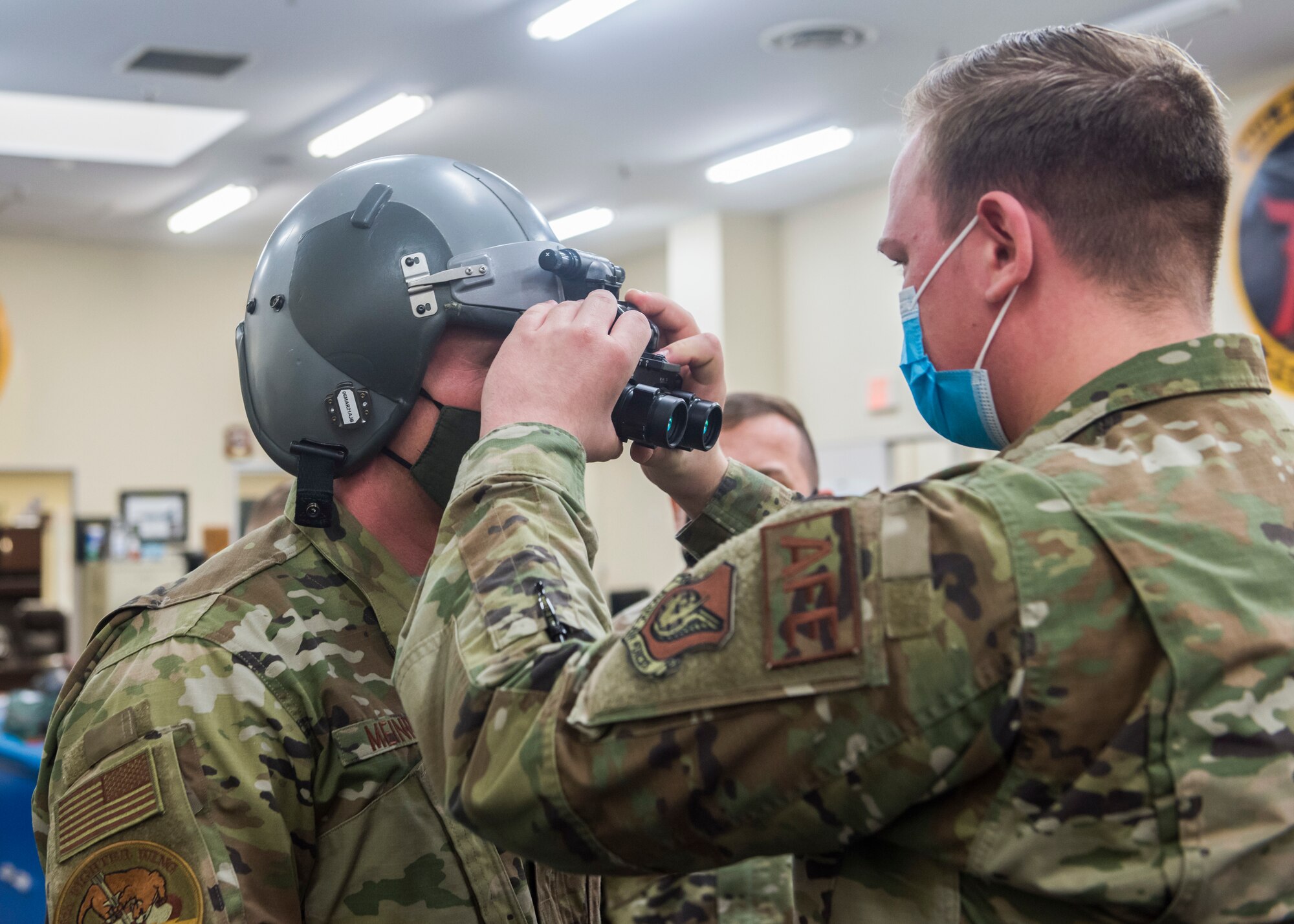 Man in uniform assist another man in uniform wearing a helmet, attach night vision goggles to it.