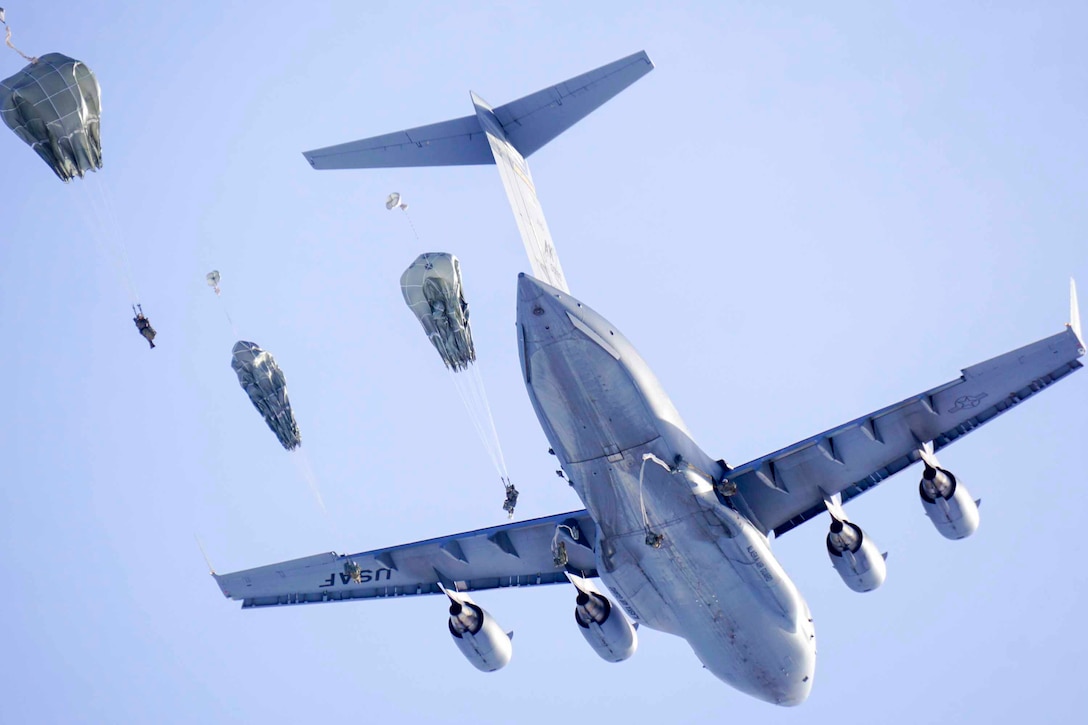 Soldiers wearing parachutes descend in the sky as a plane flies behind.