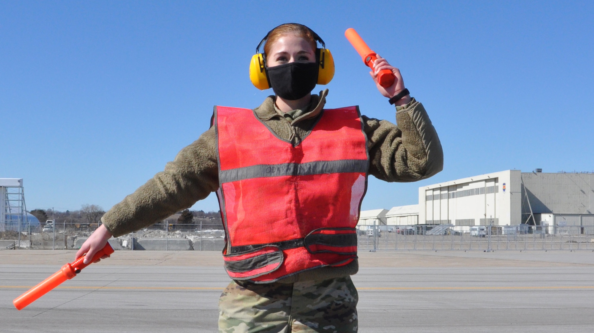 Air Force female wearing COVID-19 protective mask, ear protection headphones, in uniform, wearing an orange vest and carrying an orange signal lights in each hand directing an aircraft