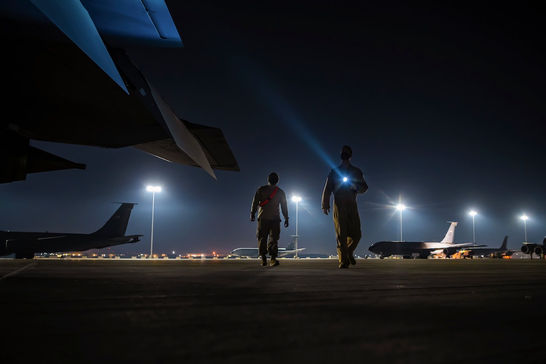 Two service members use a flashlight to walk around an aircraft in the dark.