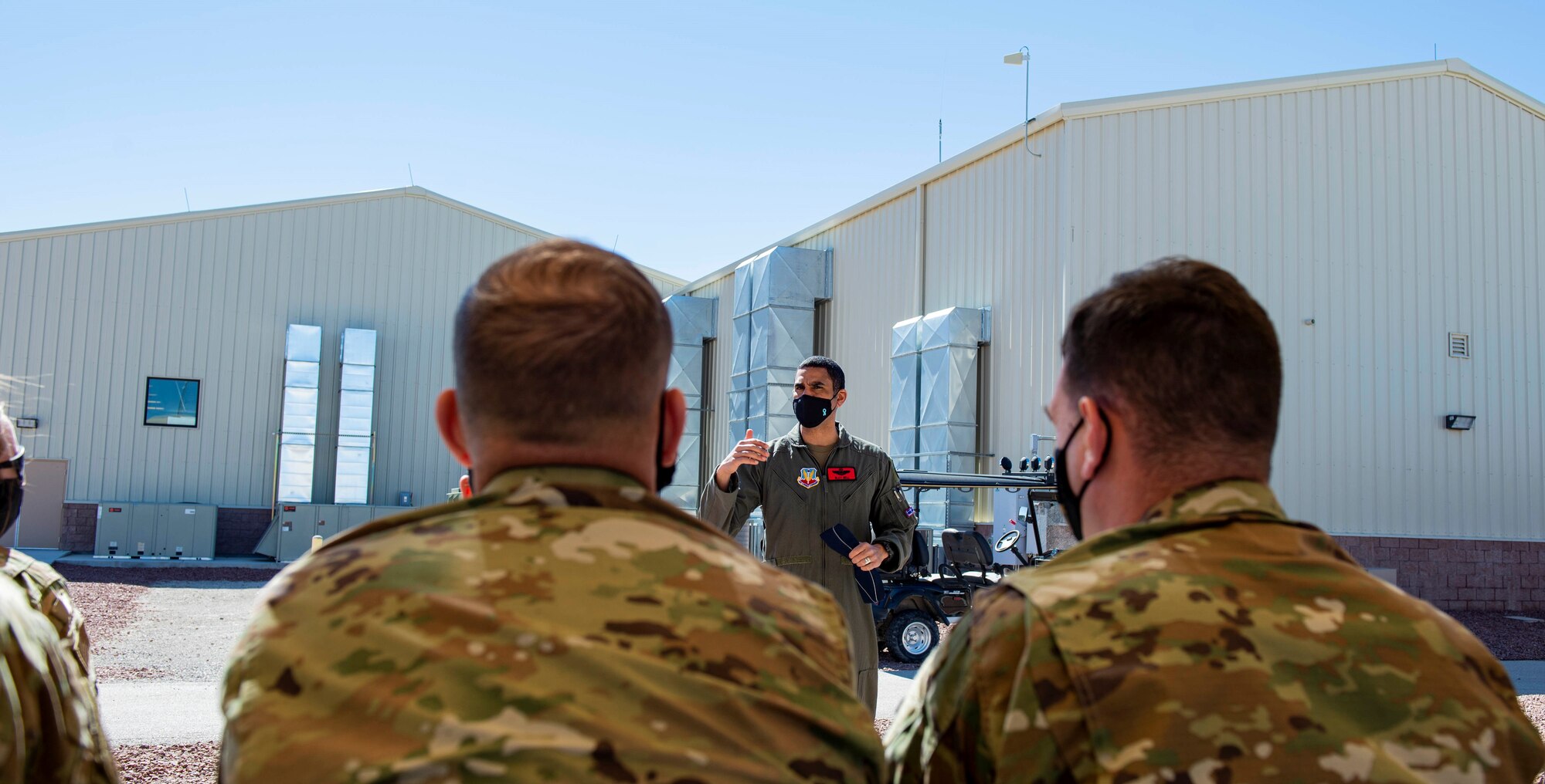 Male Colonel speaks to a group of military members outside near a picnic table area.