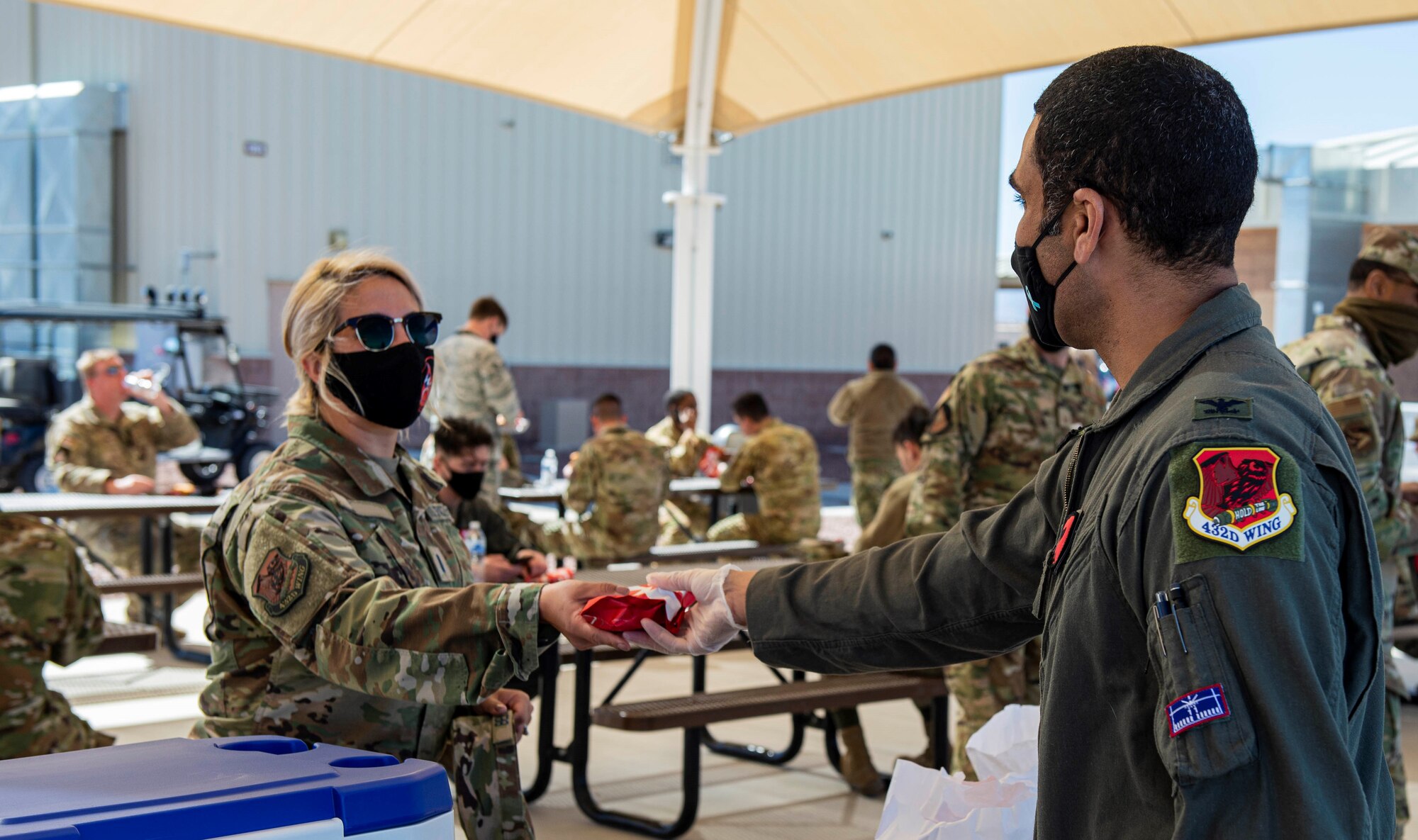 Male Colonel hands out sandwiches to a female military members standing in line outside near a park bench seating area.