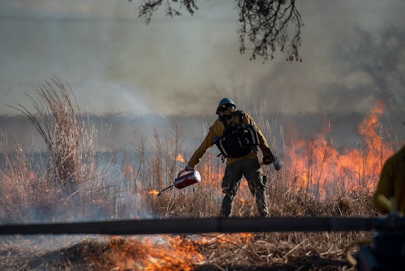 Air Force Wildland Fire Center member using torch to light grass on fire.