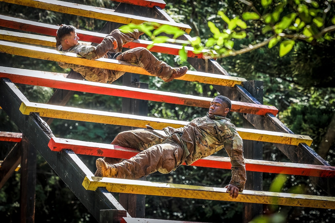 Two soldiers navigate a slanted, ladder-like obstacle in wooded surroundings.