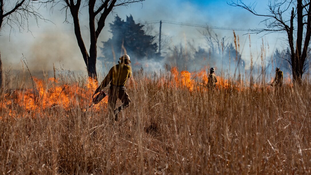 Air Force personnel use torches to set a dry grassy field on fire.