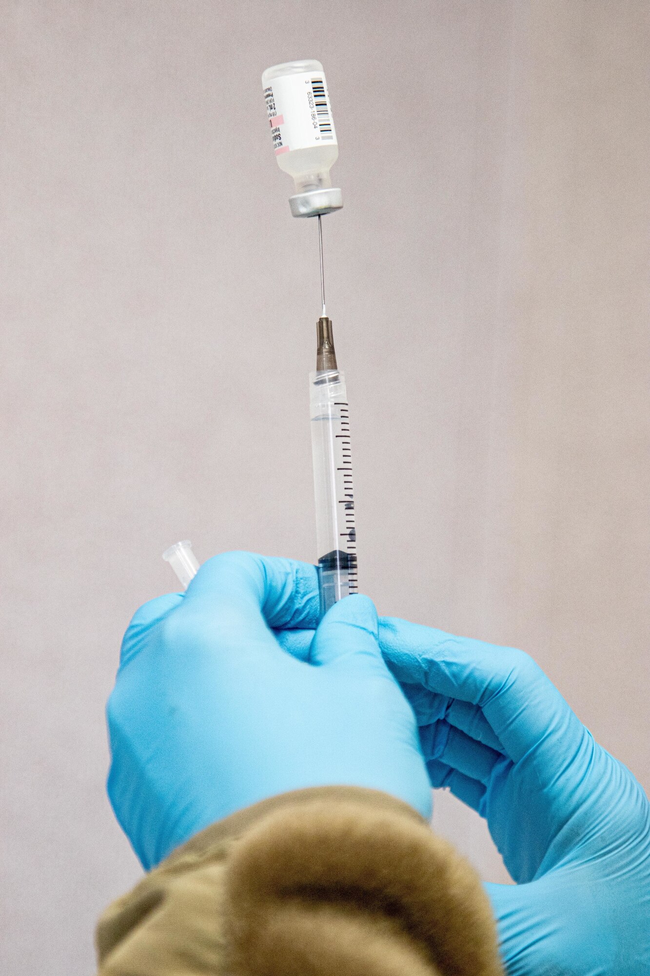 A gloved-hand holds a syringe.  Atop the syringe is a vial of vaccine.