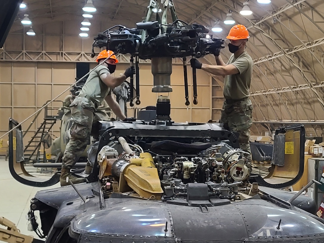 Two soldiers in camo uniforms and orange hard hats stand on top of a helicopter part to perform maintenance.