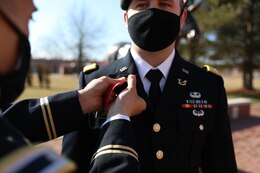 Male soldier in dress uniform pins branch insignia on male soldier in dress uniform.