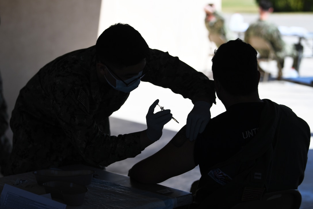 A sailor wearing a face mask and gloves gives a COVID-19 vaccine to another sailor.