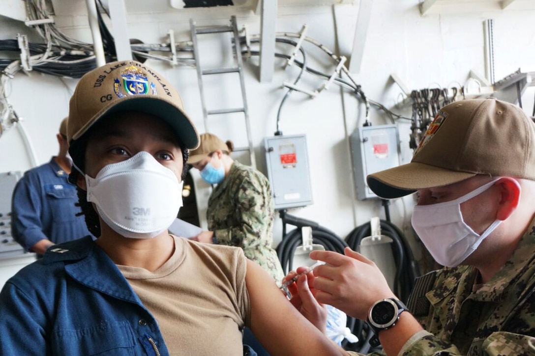 A sailor wearing a face mask gives a COVID-19 vaccine to another sailor wearing a face mask.