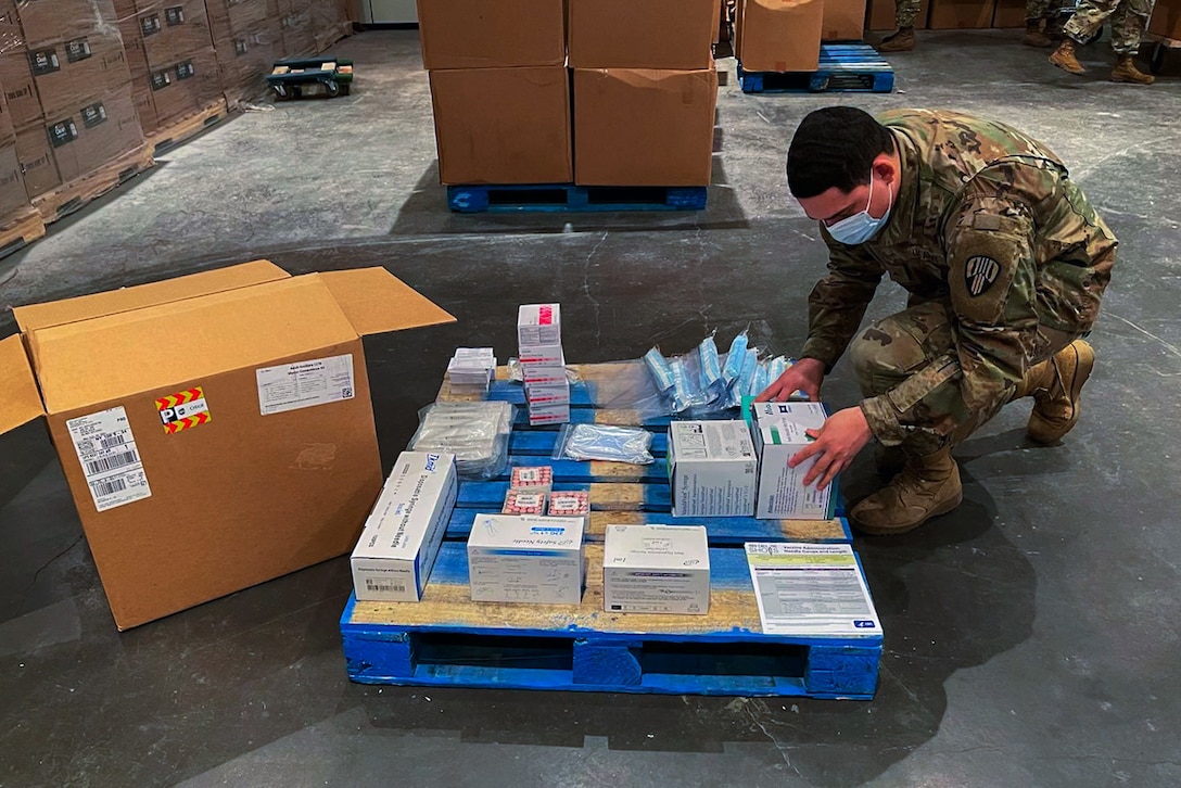 A soldier wearing a face mask kneels on a warehouse floor to arrange boxes on a pallet.