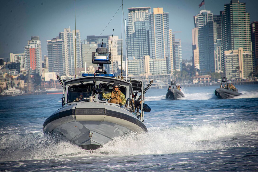 A sailor mans a machine gun in a small boat; two other boats travel behind it.