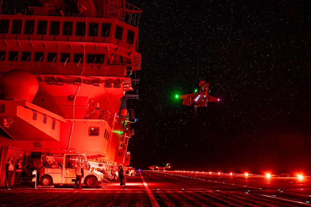 An aircraft illuminated by red light prepares to land on the deck of a ship.