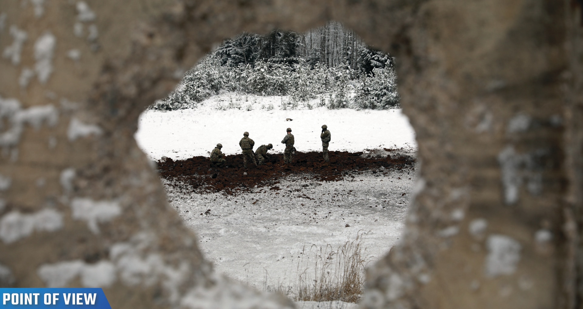 Soldiers seen through a hole in a cement wall work in a field.
