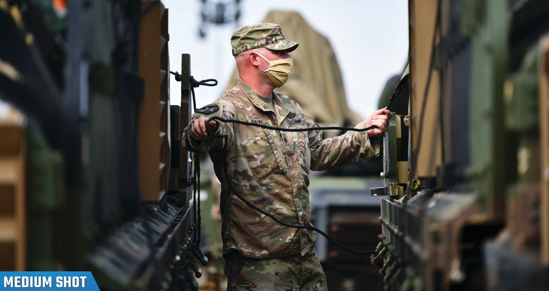 A soldier wearing a mask stands between two vehicles holding a cable.