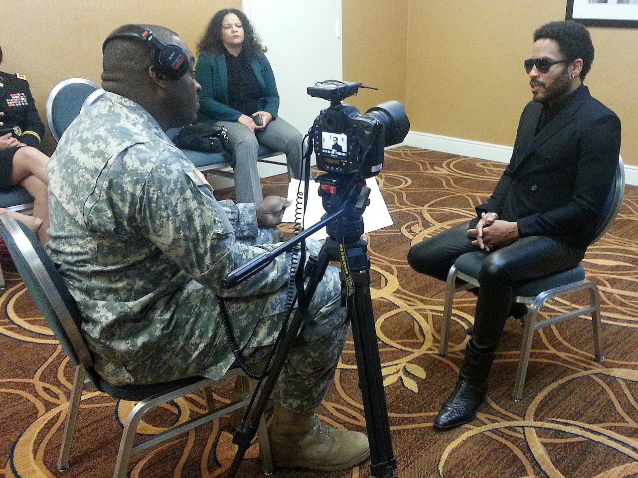 A seated man in sunglasses is filmed by a camera while talking with a soldier.