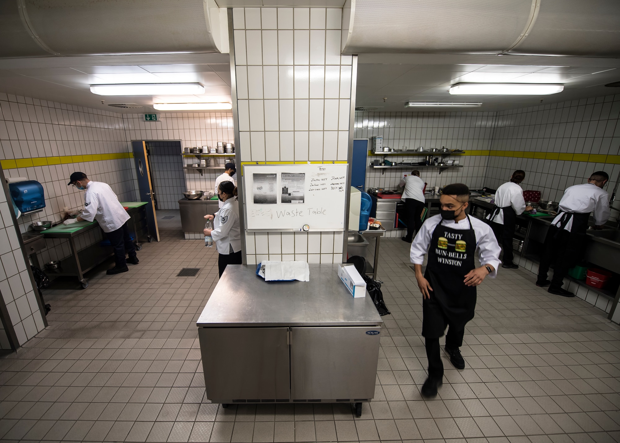 People prepare to cook in two side-by-side rooms. A man is quickly walking out of one of the rooms.
