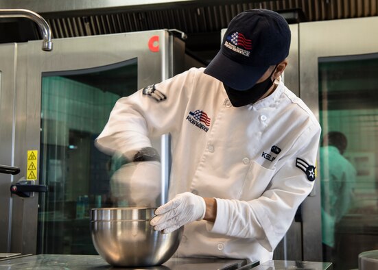 People prepare to cook in two side-by-side rooms. A man is quickly walking out of one of the rooms.