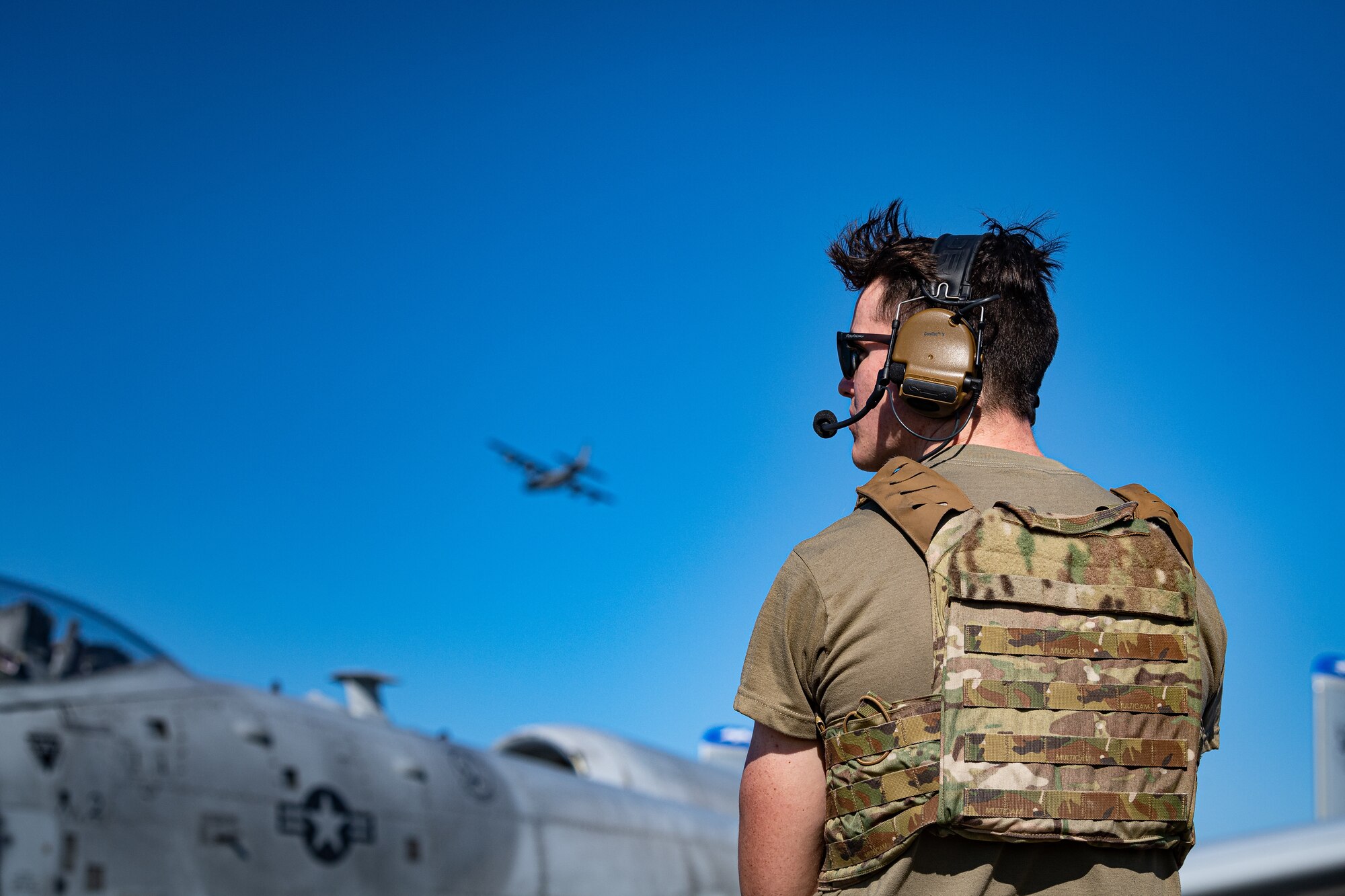 An Airman stands in front of an aircraft while another aircraft flies overhead