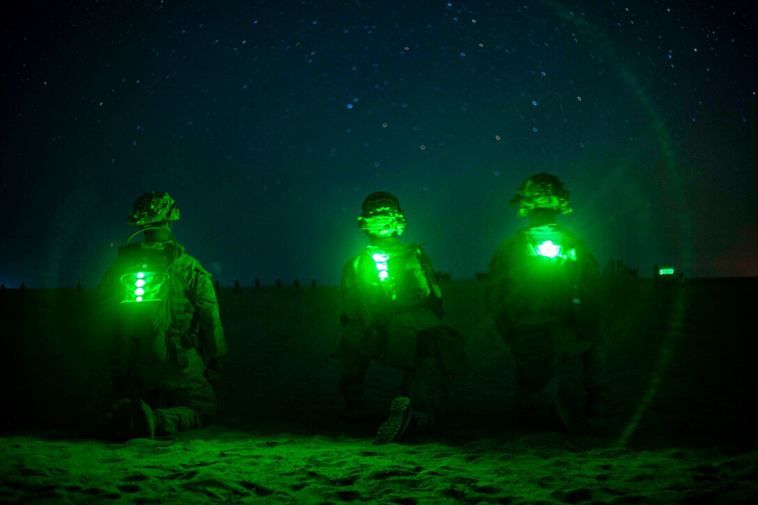 Three Marines kneel on the ground with green lights strapped to their backs at night.