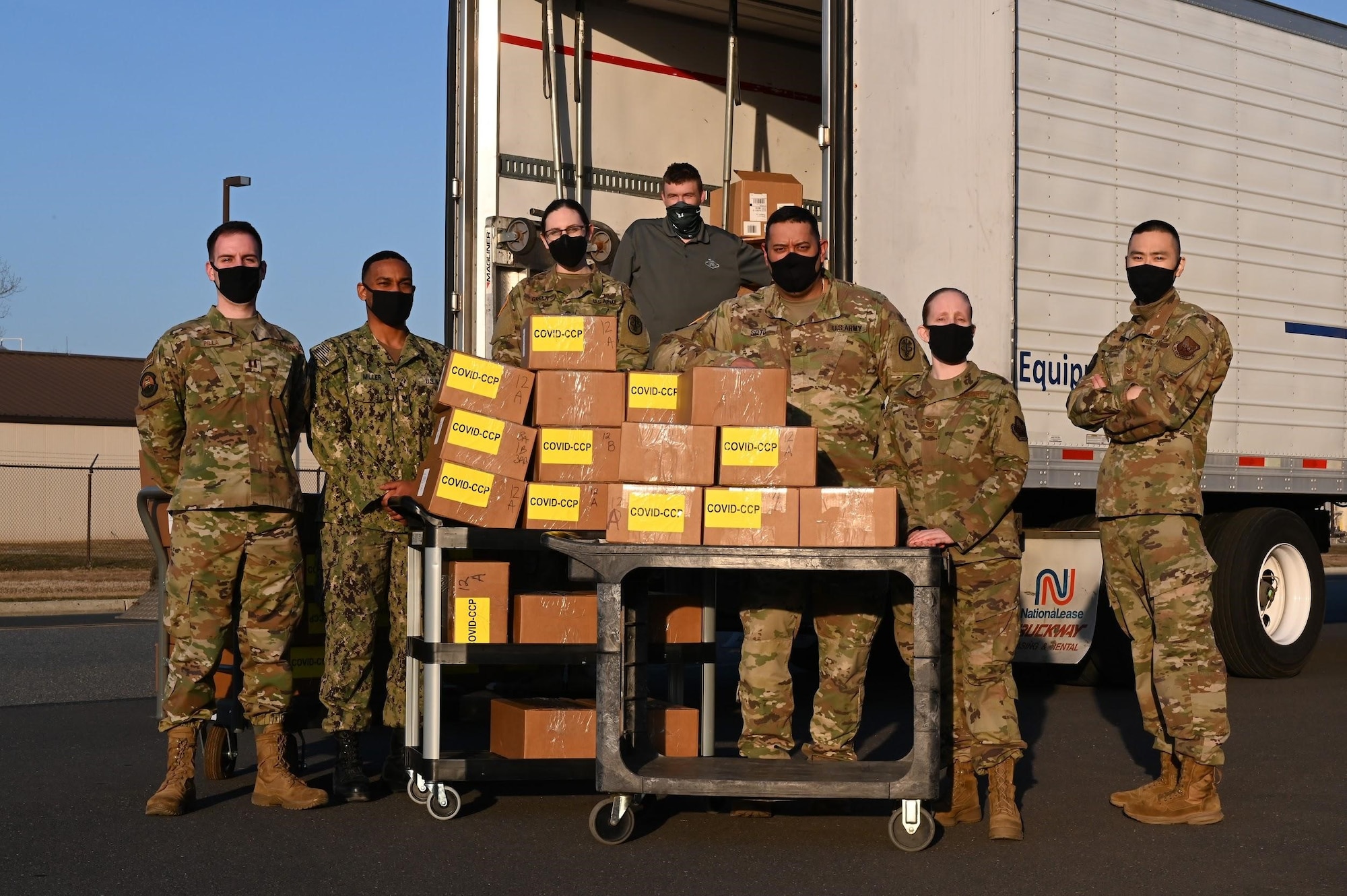 A group of seven people stand in front of a pile of boxes labeled "COVID CCP," with a delivery truck within the background