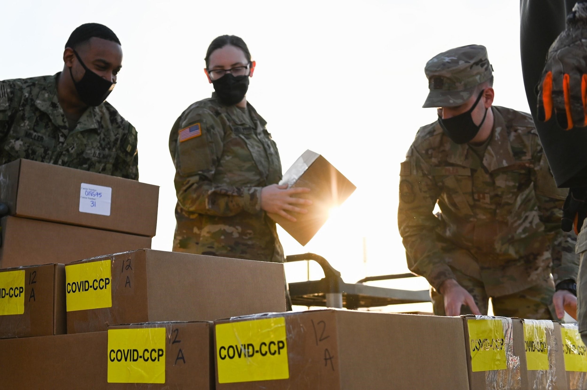 A group of service members hold boxes prepared to stack on top of an organized pile of boxes labeled "COVID - CCP"