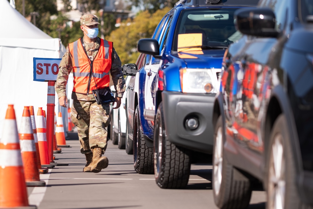 A soldier wearing a face mask walks beside a line of cars.