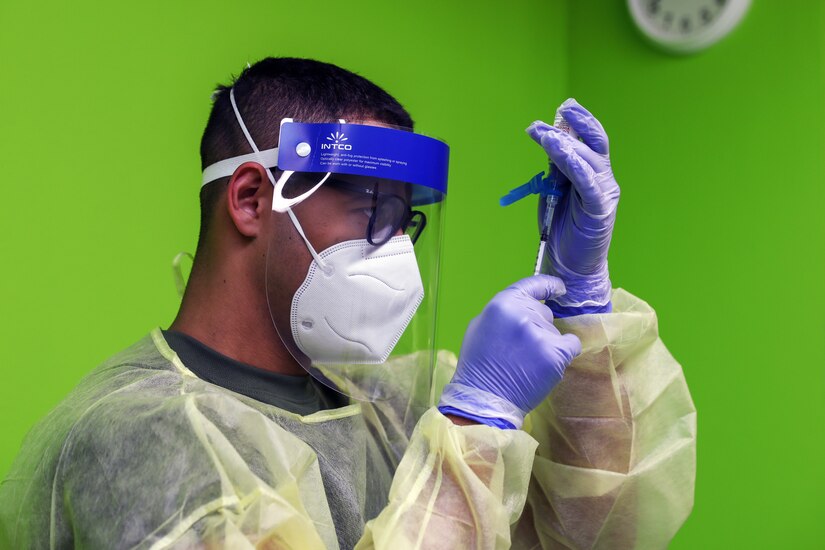 A soldier wearing personal protective equipment holds a syringe.